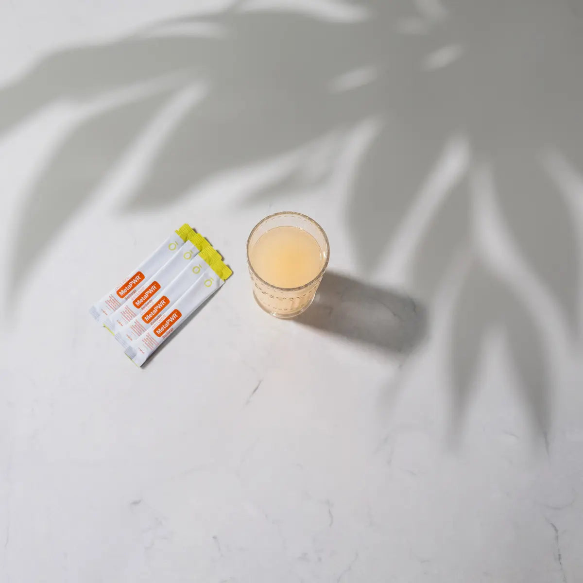 MetaPWR™ Advantage dōTERRA — sobres y vaso naranja sobre mármol con palmera al fondo