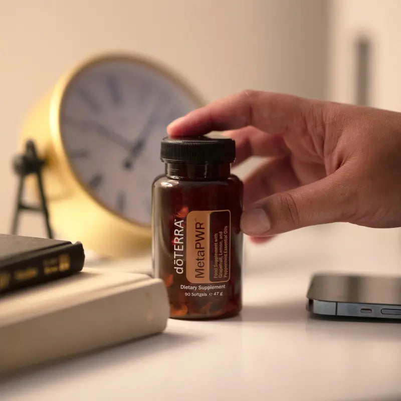 Cápsulas blandas MetaPWR™ dōTERRA — mano sosteniendo el frasco en escritorio con reloj dorado