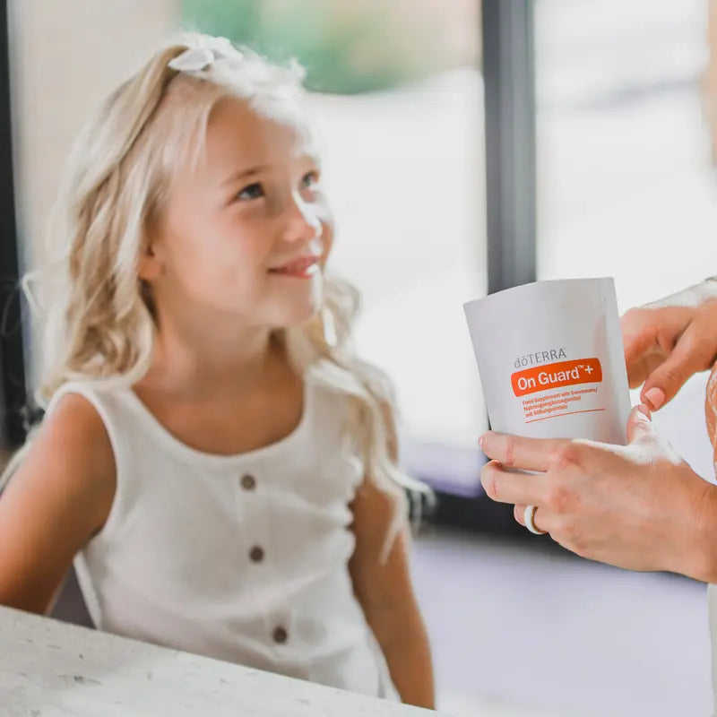 Comprimidos masticables On Guard™+ dōTERRA con niña rubia sosteniendo la bolsa en escena familiar