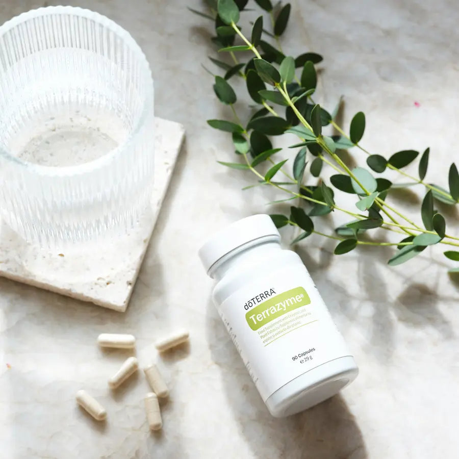 Frasco de TerraZyme dōTERRA 90ct sobre mármol con vaso de agua y eucalipto