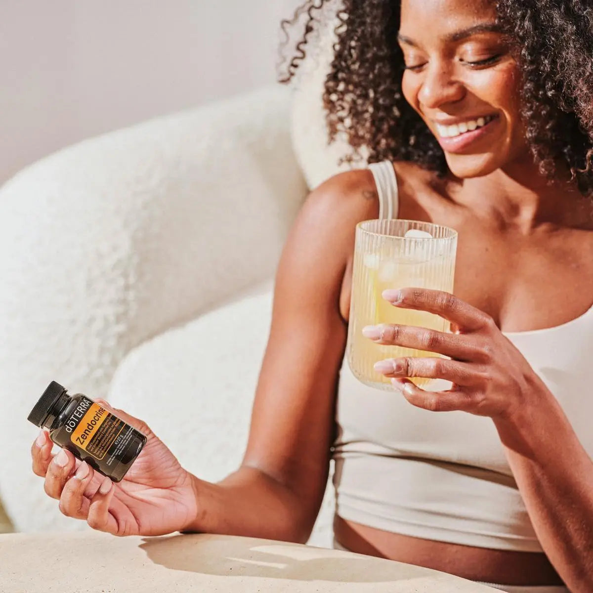 Mujer de cabello rizado con frasco negro de Zendocrine Softgels dōTERRA y vaso en el sofá