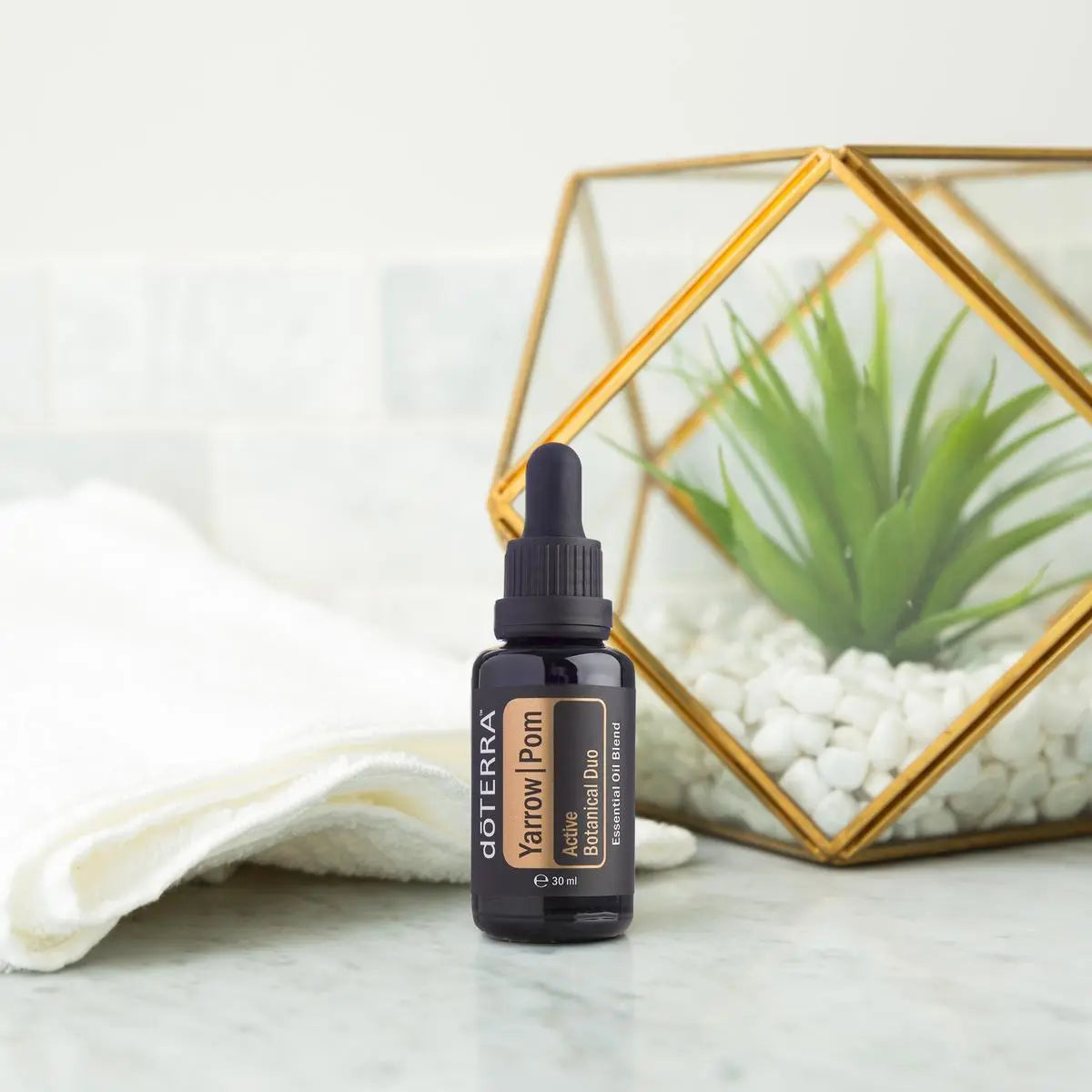 Frasco del Yarrow|Pom dōTERRA 30ml sobre superficie de mármol blanco junto a toalla enrollada blanca y terrario geométrico de latón dorado con suculenta y piedras blancas