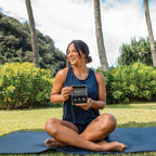 Mujer sentada en posicion de loto sobre esterilla de yoga en prado de Hawaii con palmeras, sosteniendo abierto el kit Yoga Collection doTERRA con sus aceites esenciales