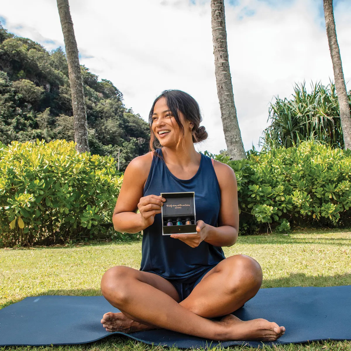 Mujer sentada en posicion de loto sobre esterilla de yoga en prado de Hawaii con palmeras, sosteniendo abierto el kit Yoga Collection doTERRA con sus aceites esenciales